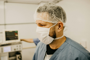 A man wears a white hairnet and mask with blue-grey scrubs in a hospital room