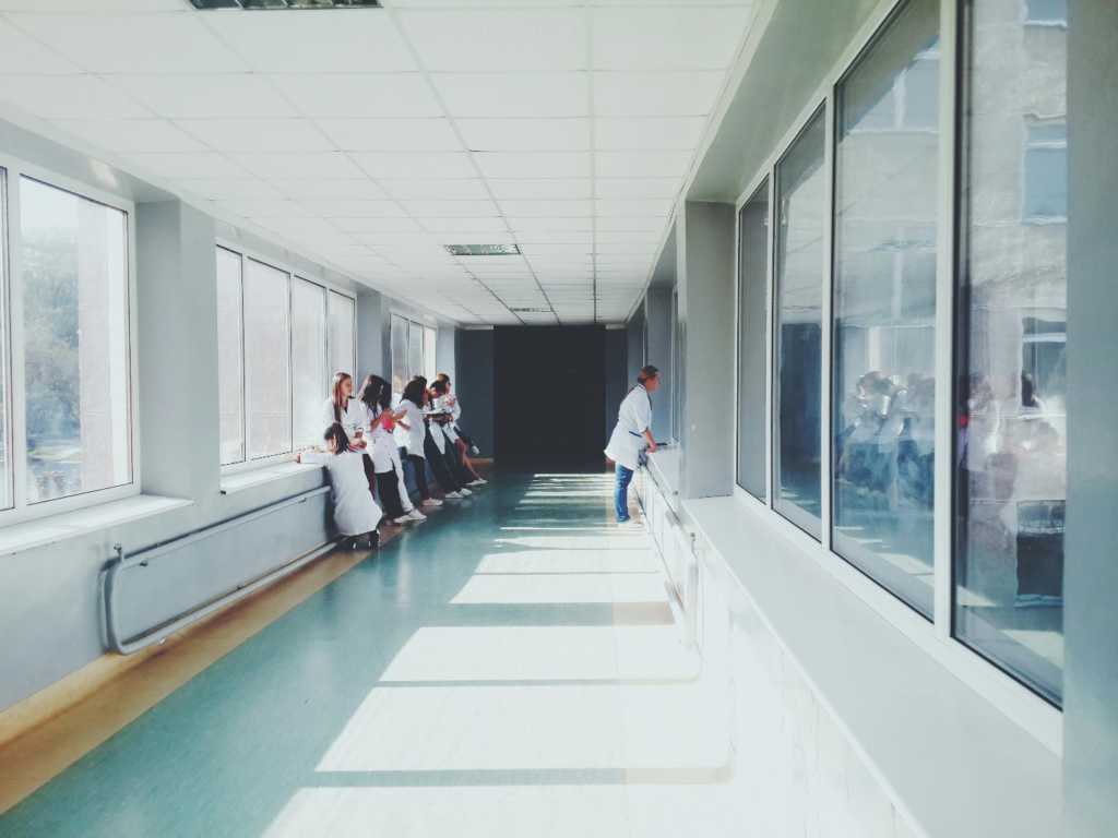 A number of people in white labcoats lean against a wall with windows in a hallway while another looks into a room