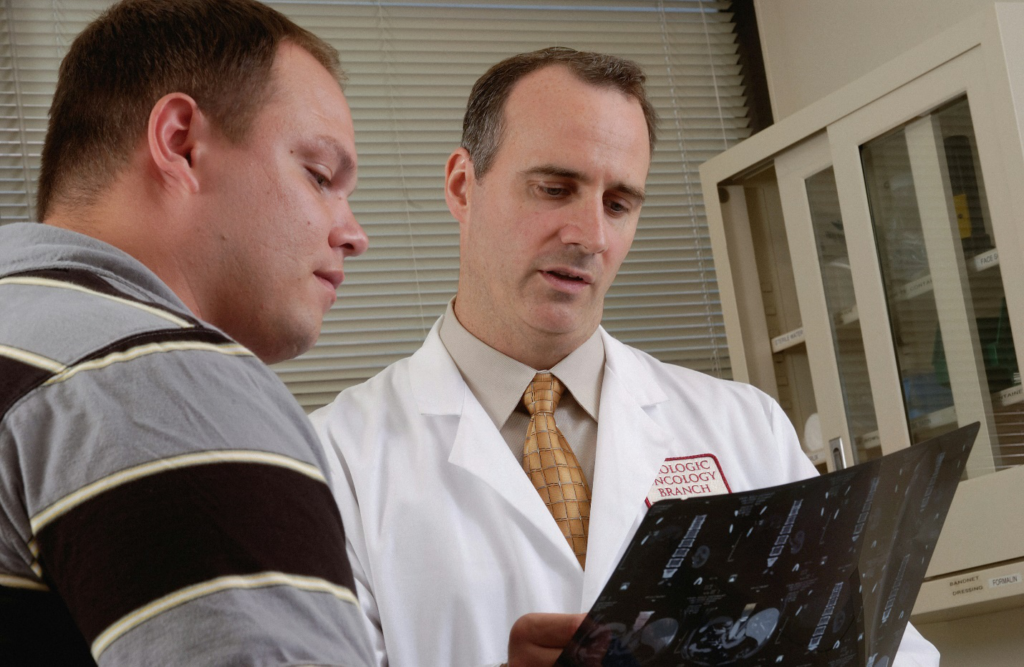 A person wearing a white lab coat holding an imaging film beside another person in a clinical setting appears to be carefully explaining test results to avoid both medical malpractice and doctor's error.