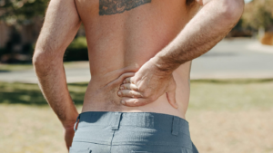 A man with a tattoo on his upper back holds his lower back while wearing grey lowers outdoors, showing how a seemingly small injury can cause lifelong pain and highlighting the need for a personal injury lawyer