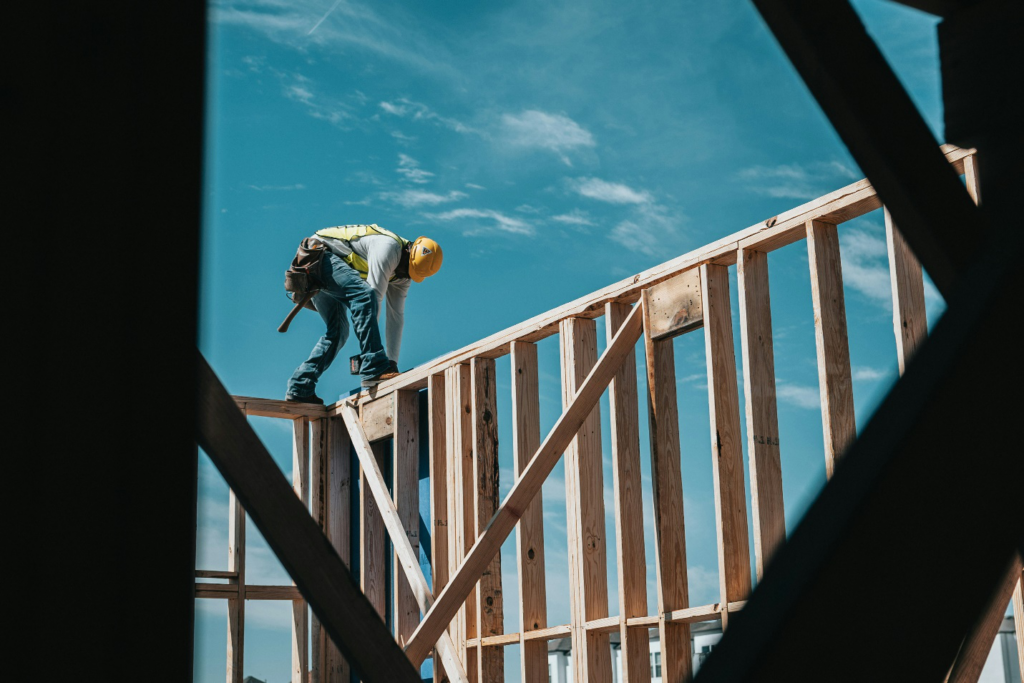 A man in a yellow hard hat standing atop a wooden frame against a bright blue sky with small scattered clouds, highlighting the precarious situation some employees work in and representing the need for workers' compensation and personal injury lawyers.