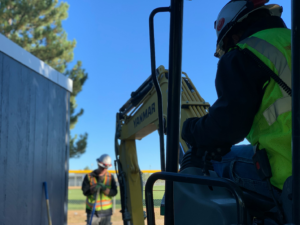 A man in a neon yellow reflective vest and white hard hat operating heavy machinery by YANMAR while another worker stands ahead outdoors, representing the importance of workers' compensation and personal injury lawyers for those who put themselves in danger on a daily basis.