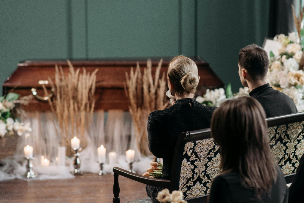 A couple sits on a bench at a funeral with a closed casket, flowers, and candles, highlighting the need for compassionate legal support for wrongful death at such a difficult time.