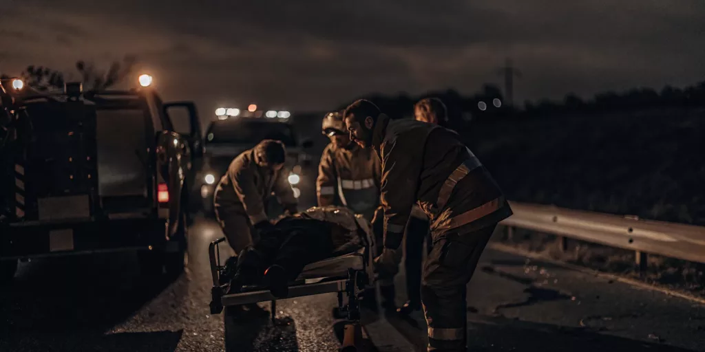 A tow truck and emergency responders at a highway crash scene at dusk helping an injured person